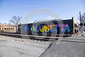A colorful wall mural with the word âMariettaâ along the railroad tracks with people standing on the corner in Marietta